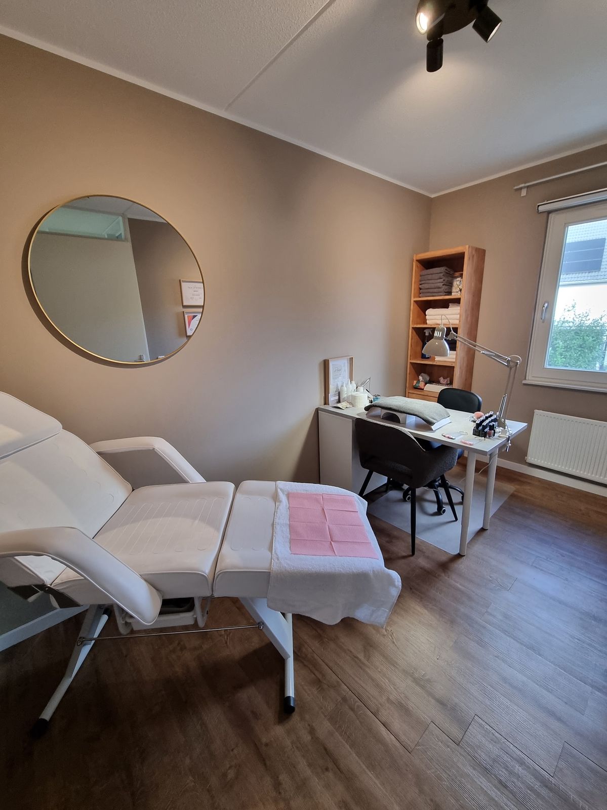 De behandelkamer met witte behandelstoel, nageltafel, ronde spiegel en taupe muren